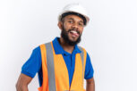 joyful young afro-american builder man in uniform with safety helmet stucks out his tongue isolated on white background with copy space