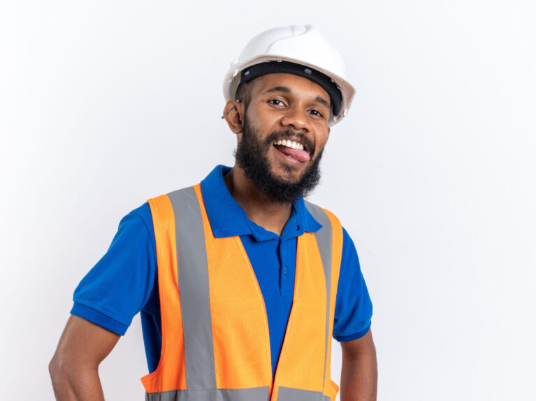 joyful young afro-american builder man in uniform with safety helmet stucks out his tongue isolated on white background with copy space