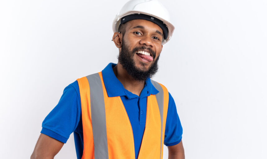 joyful young afro-american builder man in uniform with safety helmet stucks out his tongue isolated on white background with copy space